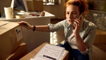 A courier looking concerned while talking on a mobile phone next to stacked delivery boxes.